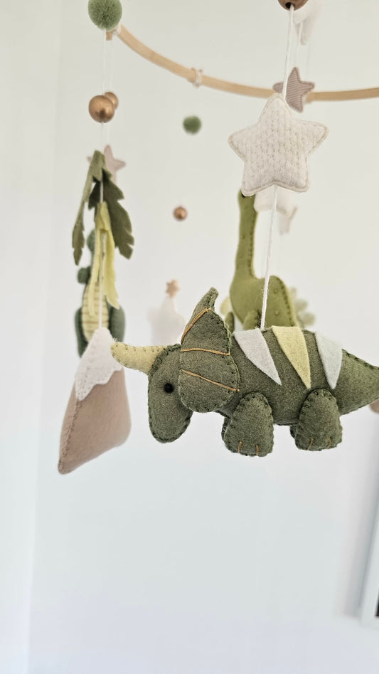 A fabric dinosaur baby mobile hanging from a wooden rod against a white wall. The mobile features an olive green felt dinosaur with cream and yellow accents, a beige pillow, green and cream pom-poms, and knitted cream stars.