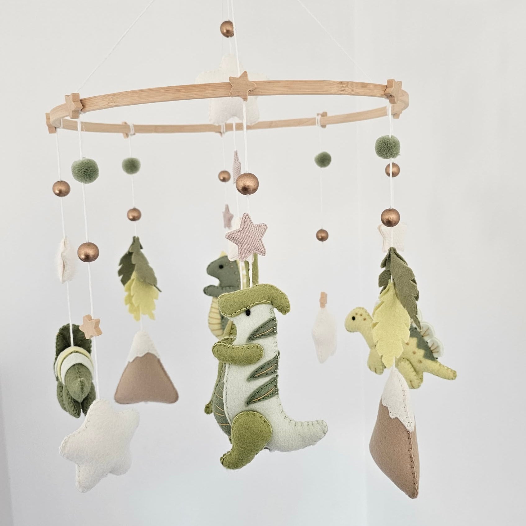 A fabric baby mobile hanging from a light wood ring, featuring green dinosaur plush figures, star and pom-pom accents, and small mountain shapes against a white wall.