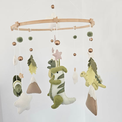 A fabric baby mobile hanging from a light wood ring, featuring green dinosaur plush figures, star and pom-pom accents, and small mountain shapes against a white wall.