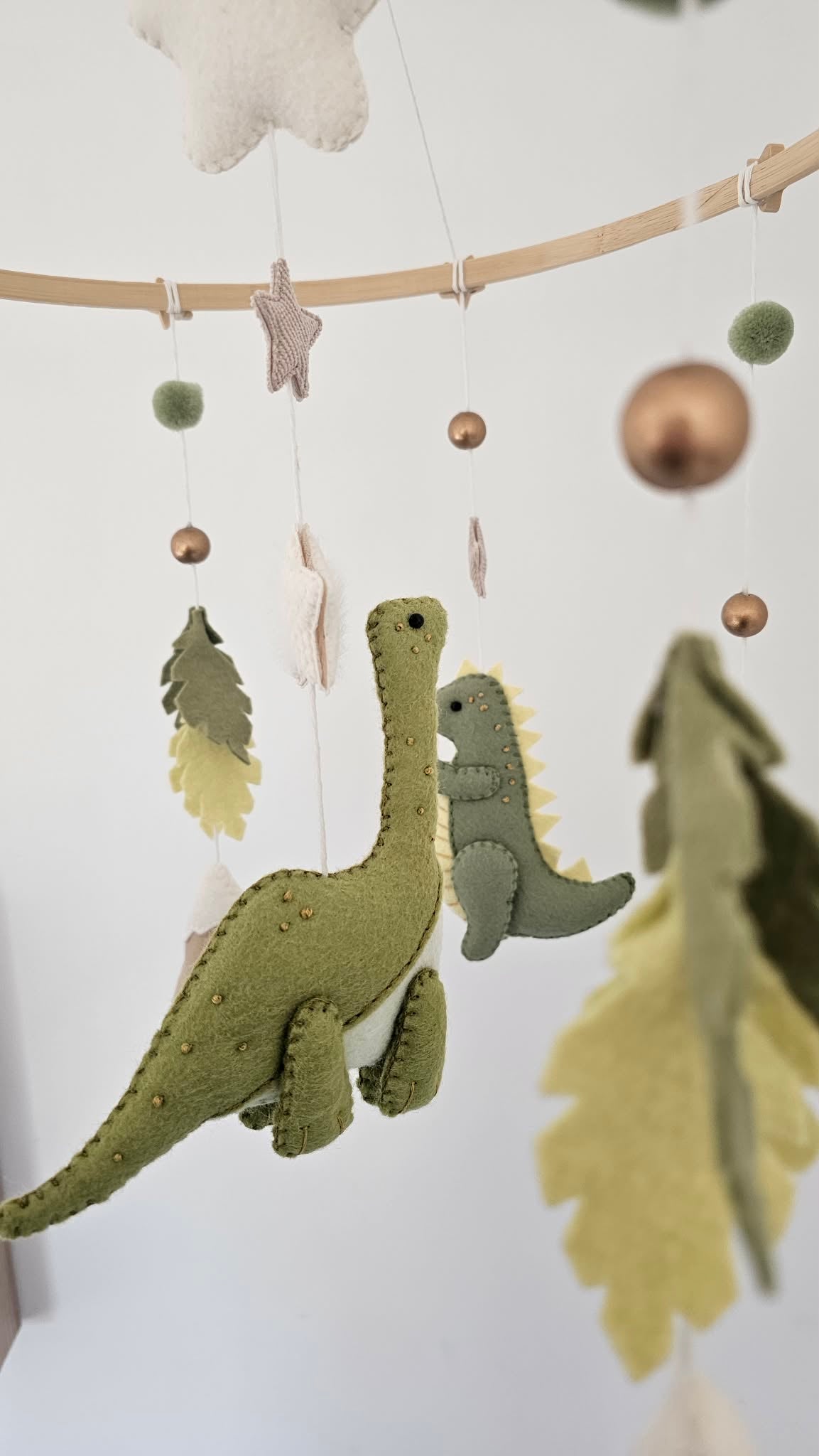 A felt baby mobile hanging from a wooden rod, featuring two green dinosaurs, leaf shapes, pom-poms, and metallic gold and green balls against a soft white background.