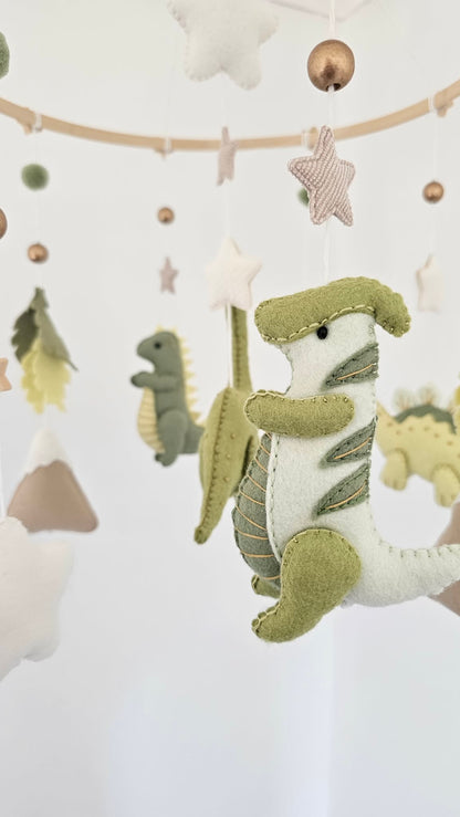 A felt baby mobile with a green and white dinosaur hanging from a wooden rod, surrounded by star, cloud, and mountain shapes in soft neutral tones.