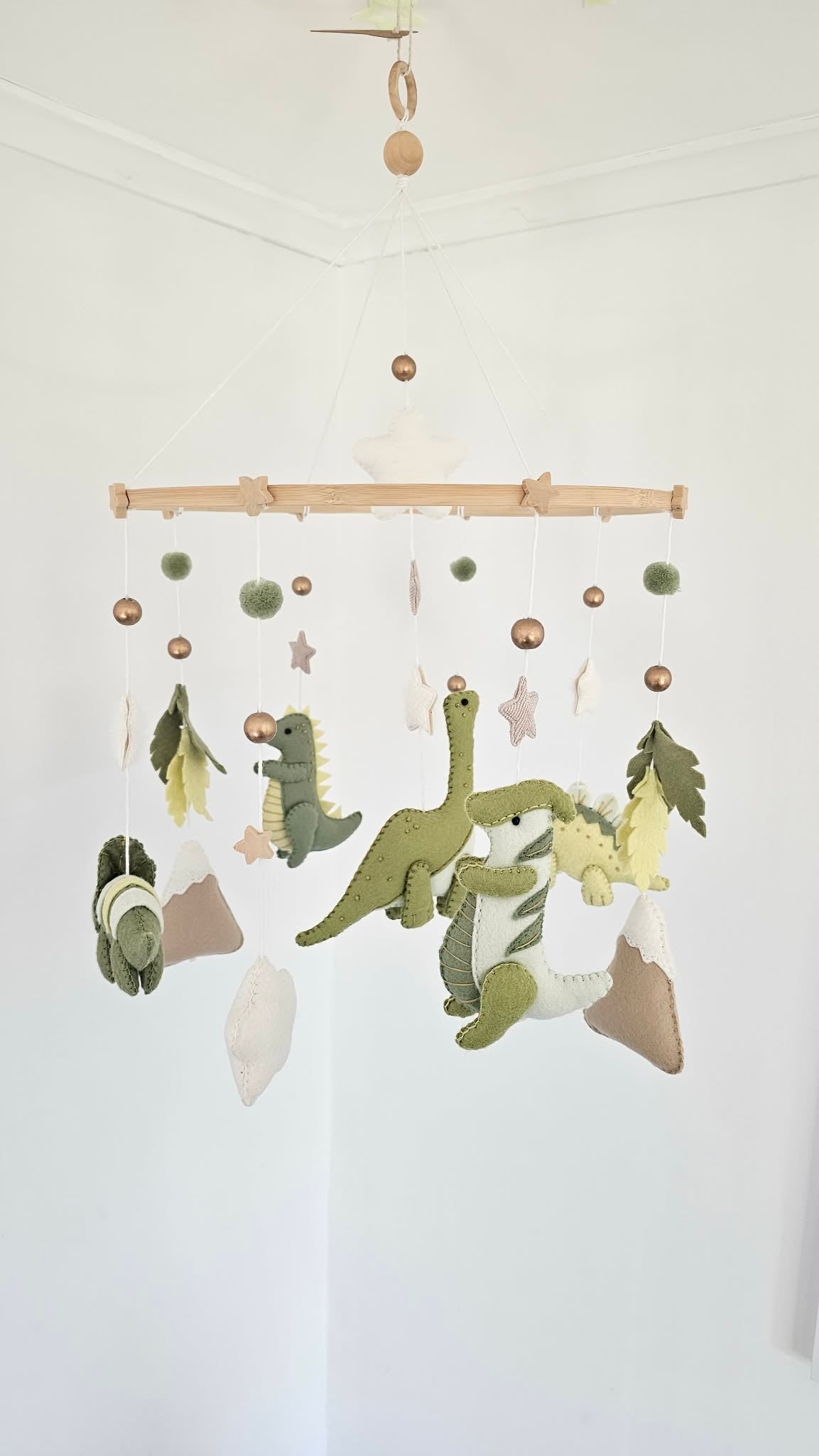 A felt dinosaur baby mobile hanging from a wooden bar in a white nursery corner. The mobile features green and yellow dinosaurs, white stars, green pom-poms, and brown and beige planets.