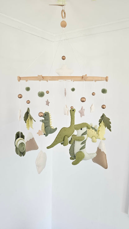 A felt dinosaur baby mobile hanging from a wooden bar in a white nursery corner. The mobile features green and yellow dinosaurs, white stars, green pom-poms, and brown and beige planets.