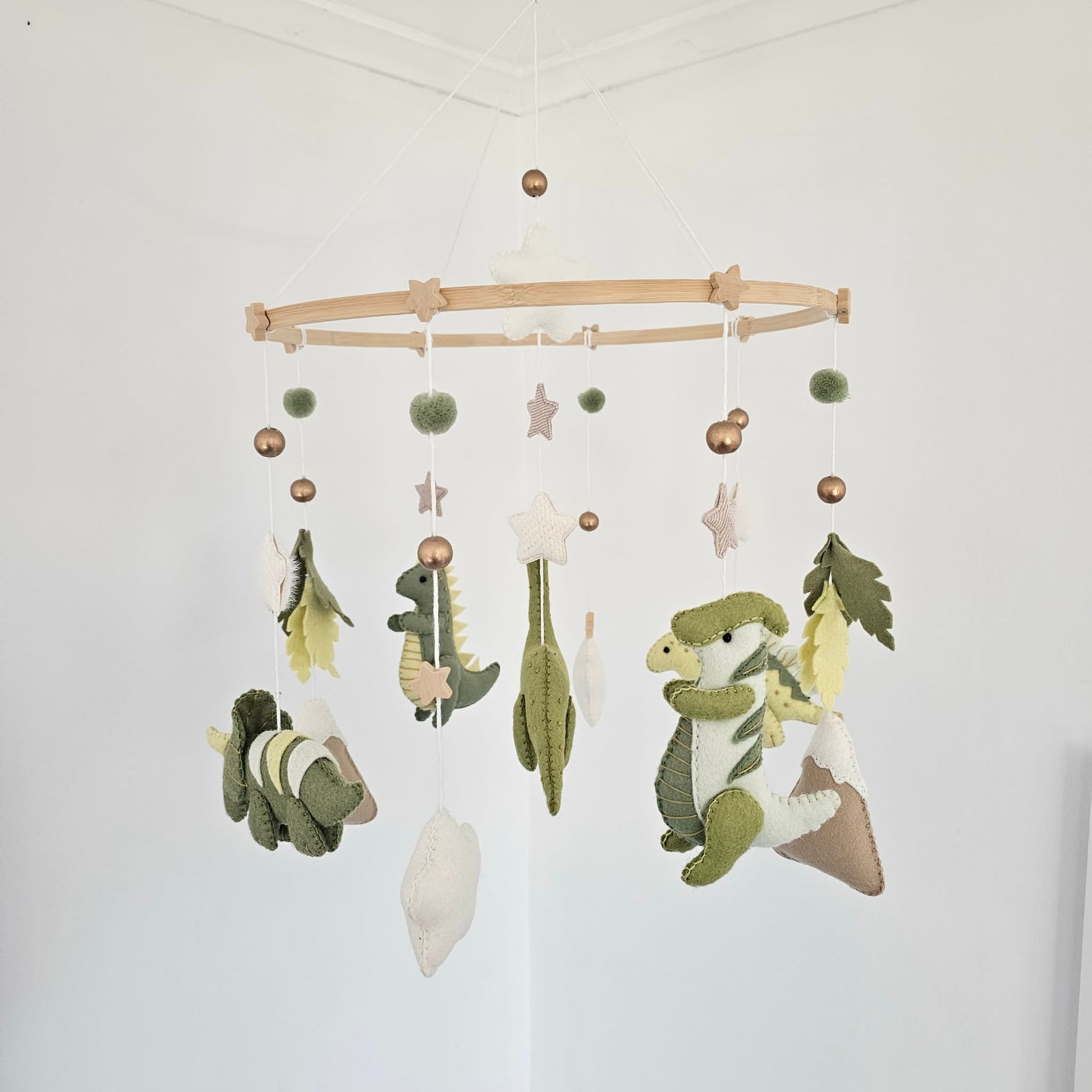 A wooden hoop baby mobile hanging from the ceiling, featuring green felt dinosaurs, cacti, leaves, stars, rabbits, and pom-poms in a soft neutral palette.