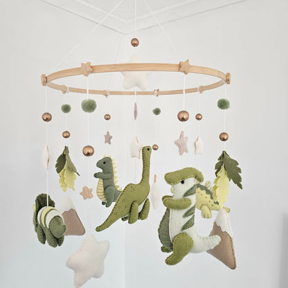 A felt dinosaur baby mobile hanging from a wooden hoop. The mobile features plush green and white dinosaurs, star shapes, pom-poms, and leaf accents in muted sage, olive, cream, and metallic rose-gold tones, suspended against a plain white wall.