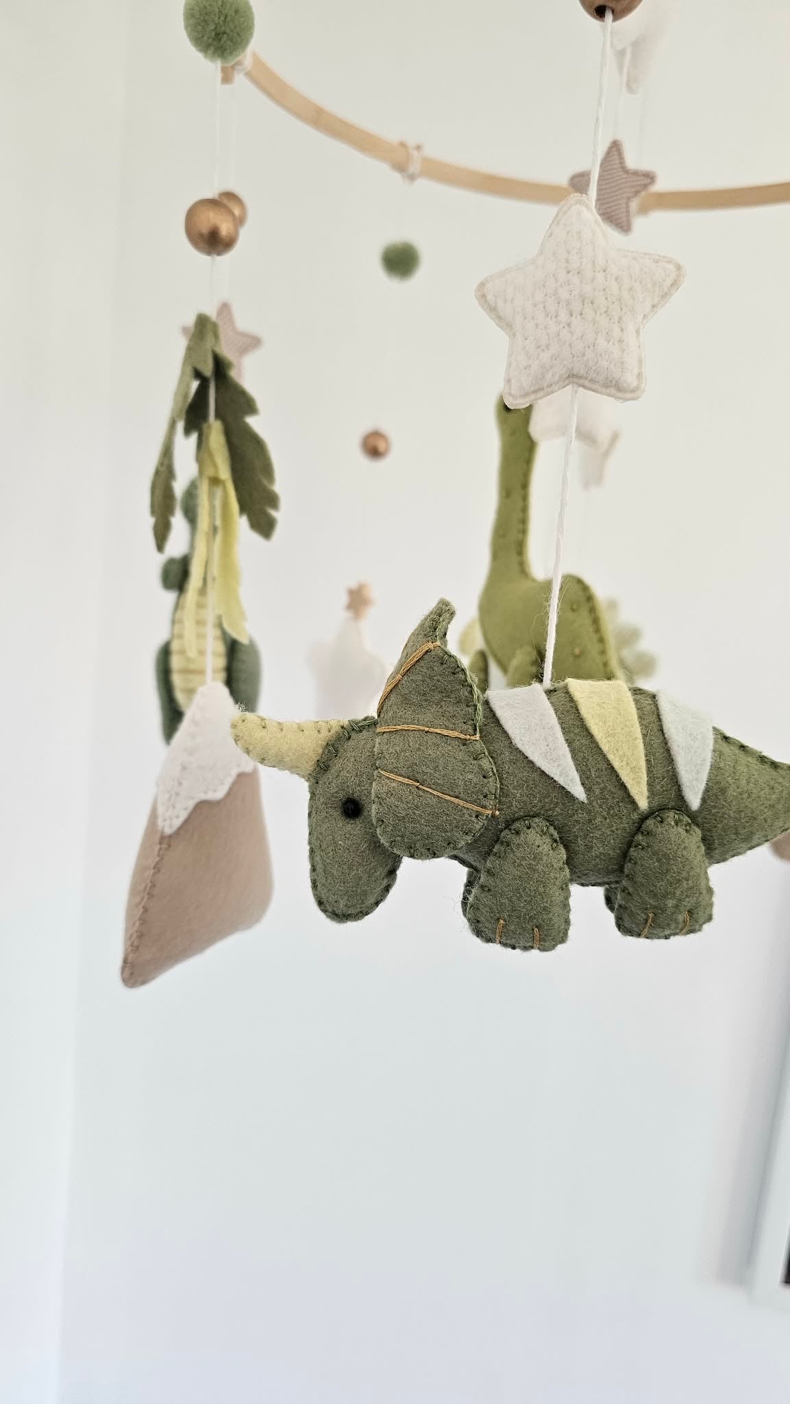 A fabric dinosaur baby mobile hanging from a wooden rod against a white wall. The mobile features an olive green felt dinosaur with cream and yellow accents, a beige pillow, green and cream pom-poms, and knitted cream stars.